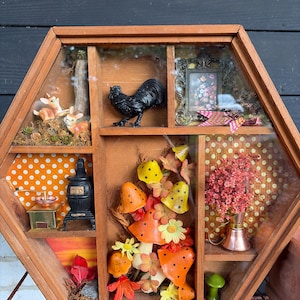 May include: A hexagonal wooden shadow box display case featuring multiple compartments. Contents include two small deer figurines, a black rooster, a framed floral picture, a miniature stove, a coffee grinder, artificial mushrooms, and flowers.