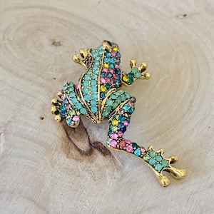 May include: A gold-toned frog brooch with a detailed design of colorful rhinestones. The frog is in a sitting position with its legs outstretched.
