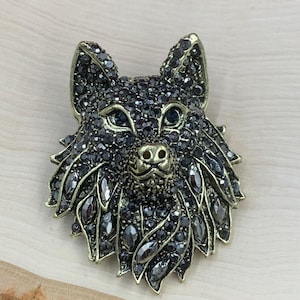 May include: A detailed brooch in the shape of a wolf's head. The brooch is primarily black with dark gray crystals and a gold-colored nose and ears. The wolf's head is set against a light wood grain background.