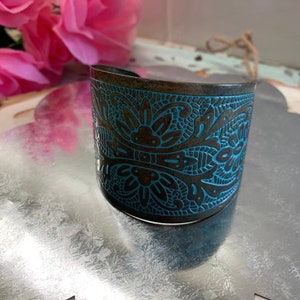 Op de afbeelding: Een brede metalen armband met een ingewikkeld blauw en bruin bloemenmotief. De armband is gemaakt van donker metaal met een licht verweerde afwerking.