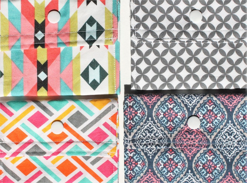 May include: Four fabric pouches with different patterns, each with a buttonhole and a white button. The pouches are arranged in two rows of two. The top row features a geometric pattern in pink, blue, and yellow, and a gray and white pattern. The bottom row features a geometric pattern in pink, blue, and orange, and a blue and pink pattern.