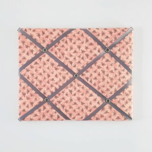 May include: A pink fabric notice board with grey ribbon crisscrossing the surface. The board has five silver push pins.