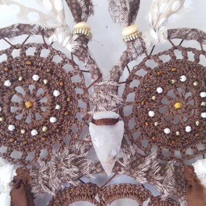 May include: A brown crocheted owl dreamcatcher with white beads and feathers. The owl's eyes are made of brown crocheted circles with white beads and gold accents. A clear quartz crystal is hanging from the owl's beak.