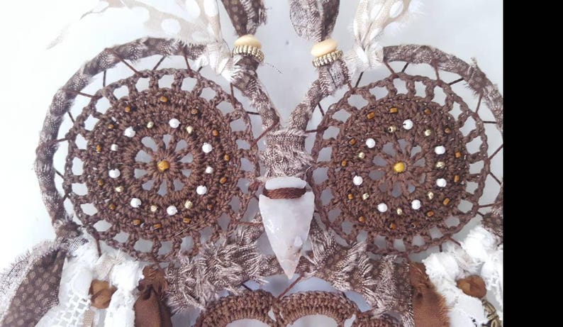 May include: A brown crocheted owl dreamcatcher with white beads and feathers. The owl's eyes are made of brown crocheted circles with white beads and gold accents. A clear quartz crystal is hanging from the owl's beak.