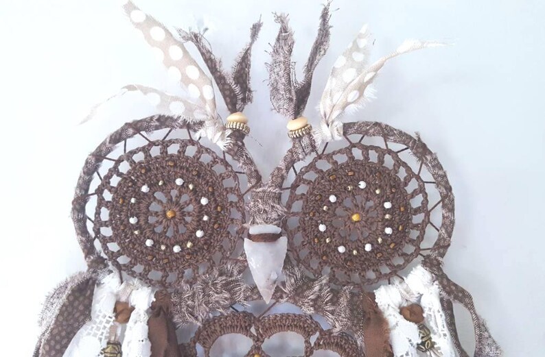 May include: A brown crocheted owl dreamcatcher with white feathers and beads. The owl's eyes are made of brown crocheted circles with white beads. The owl's beak is made of a white stone.