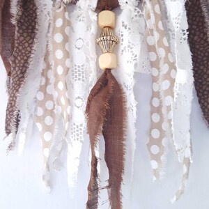 May include: A brown and white dream catcher with a beaded tassel. The dream catcher has a crocheted circle with brown and white fabric strips hanging from it. The beaded tassel is made of wooden beads and a gold metal bead.