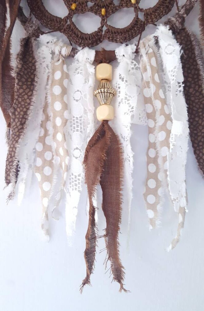 May include: A brown and white dream catcher with a beaded tassel. The dream catcher has a crocheted circle with brown and white fabric strips hanging from it. The beaded tassel is made of wooden beads and a gold metal bead.