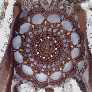 May include: A brown crocheted dreamcatcher with white and yellow beads. The dreamcatcher is surrounded by white lace and brown fabric.