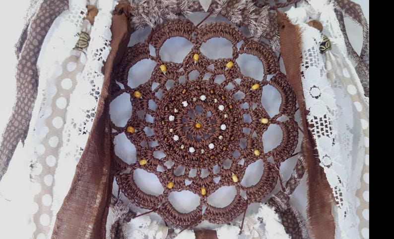 May include: A brown crocheted dreamcatcher with white and yellow beads. The dreamcatcher is surrounded by white lace and brown fabric.