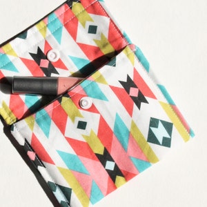 May include: A small, white fabric pouch with a geometric pattern in shades of pink, blue, yellow, and black. The pouch has two white buttons and is open to reveal a pink and gray lipstick.