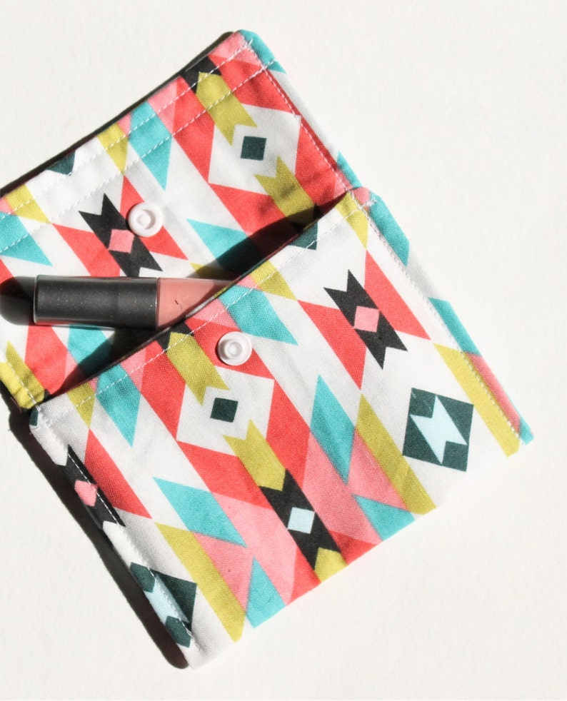 May include: A small, white fabric pouch with a geometric pattern in shades of pink, blue, yellow, and black. The pouch has two white buttons and is open to reveal a pink and gray lipstick.