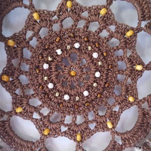 May include: A brown crocheted dreamcatcher with white and gold beads. The dreamcatcher has a circular design with a central circle of beads.