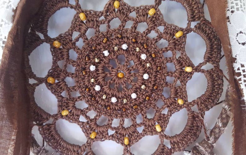 May include: A brown crocheted dreamcatcher with white and gold beads. The dreamcatcher has a circular design with a central circle of beads.