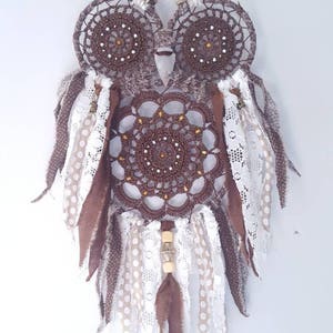May include: A brown crocheted owl dreamcatcher with white and brown feathers, white lace, and brown and white polka dot fabric. The dreamcatcher has a wooden bead and a metal bead hanging from it.
