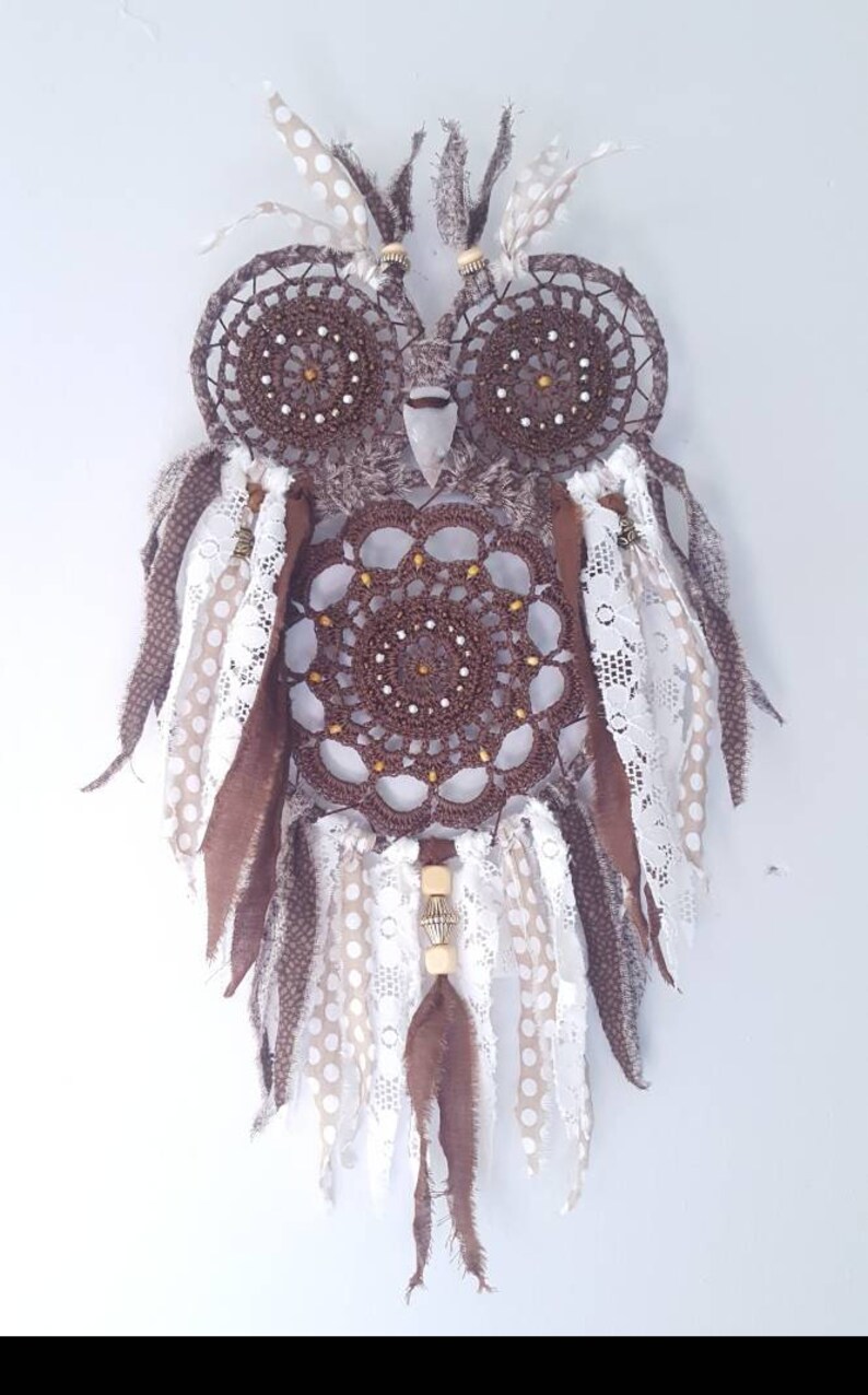 May include: A brown crocheted owl dreamcatcher with white and brown feathers, white lace, and brown and white polka dot fabric. The dreamcatcher has a wooden bead and a metal bead hanging from it.