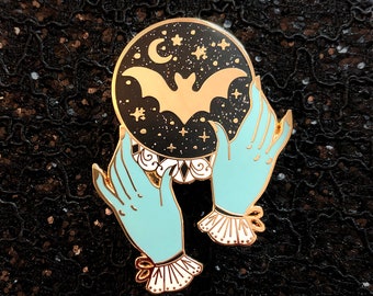 May include: Gold enamel pin featuring a pair of hands holding a black orb with a gold crescent moon, stars, and a bat inside. The hands are light blue with white cuffs and bows.