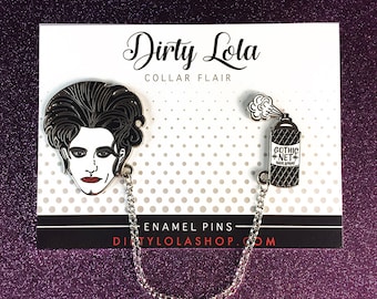 May include: Two enamel pins connected by a silver chain. The first pin features a black and white illustration of a person with a bouffant hairstyle. The second pin is a silver spray can with the text "Gothic Net Hair Spray". The pins are on a white card with the text "Dirty Lola Collar Flair Enamel Pins dirtylolashop.com".