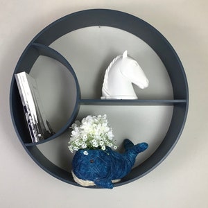 Puede incluir: Una estantería circular de pared de color azul oscuro con tres estantes. El estante superior tiene una figurita de cabeza de caballo de cerámica blanca. El estante del medio está vacío. El estante inferior tiene una maceta con forma de ballena azul con flores blancas.