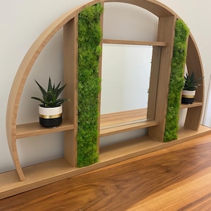 May include: A wooden, arched shelf with a central mirror and two small shelves. The shelf is decorated with green moss and holds two small potted plants. The design is modern and suitable for displaying small items.