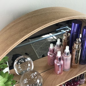 May include: A wooden shelf with a mirrored back, displaying a variety of beauty products, including glass bottles with droppers, and two glass orbs with bubbles. The shelf is made of light brown wood and has a curved shape.