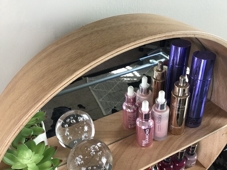 May include: A wooden shelf with a mirrored back, displaying a variety of beauty products, including glass bottles with droppers, and two glass orbs with bubbles. The shelf is made of light brown wood and has a curved shape.