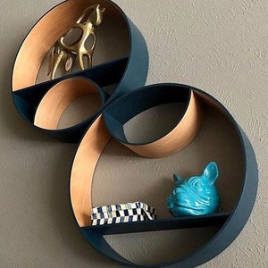 Puede incluir: Un conjunto de tres estantes circulares de pared hechos de madera y pintados de negro. Los estantes están dispuestos en un diseño único y superpuesto. El estante superior tiene una figurita de animal dorada, el estante del medio está vacío y el estante inferior tiene una figurita de animal de cerámica azul y una pequeña caja con un patrón de cuadros negros y blancos.