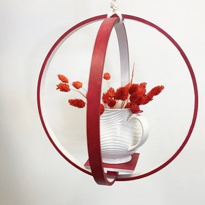 May include: A red wooden ring with a white ceramic pitcher and orange dried flowers hanging from a white rope. The ring is suspended from the ceiling.