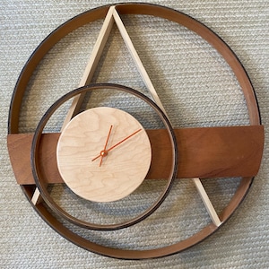Op de afbeelding: Een wandklok met een grote ronde houten lijst met een donkerbruine buitenkant en een licht houten binnenkant. Een horizontale houten plank loopt door het midden en ondersteunt een kleinere ronde licht houten wijzerplaat met feloranje wijzers.