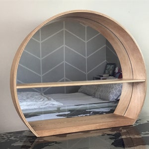 May include: A round wooden shelf with a mirror in the back. The shelf has two shelves and is mounted on the wall. The mirror reflects a bed with a white blanket and a grey patterned pillow.