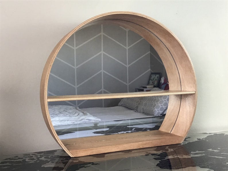 May include: A round wooden shelf with a mirror in the back. The shelf has two shelves and is mounted on the wall. The mirror reflects a bed with a white blanket and a grey patterned pillow.