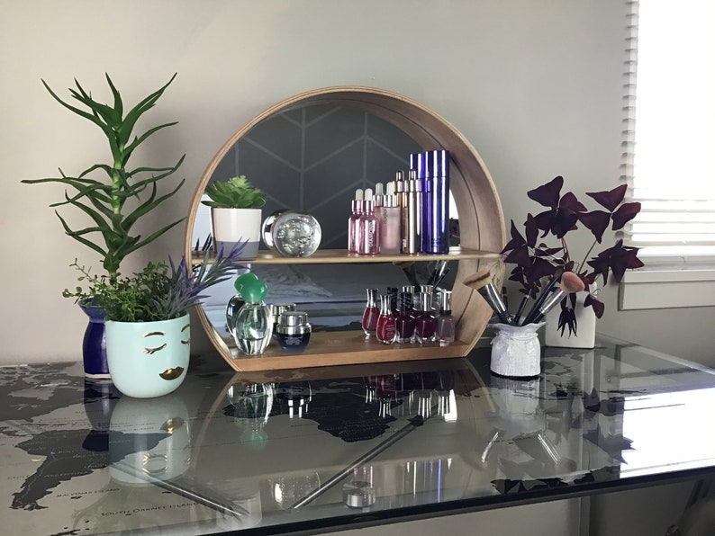 May include: A round wooden shelf with a mirrored back, displaying various bottles of cosmetics and nail polish. The shelf is on a glass table with a world map design. There are two potted plants on the table, one with green leaves and the other with purple leaves.
