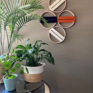 Puede incluir: Cuatro aros de bordado de madera con hilo de diferentes colores envueltos alrededor de ellos están dispuestos en forma de diamante en una pared beige. Dos de los aros tienen hilo blanco, uno tiene hilo negro y uno tiene hilo naranja. Los aros están colgados en la pared con una planta en una maceta blanca y marrón sobre una mesa de vidrio en primer plano.
