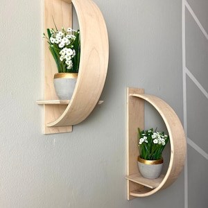 May include: Two wooden wall shelves with curved edges, each holding a small potted plant with white flowers. The shelves are mounted on a light gray wall.