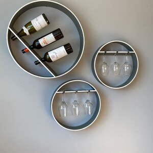 Può includere: Tre porta bottiglie da vino circolari a parete con finitura grigia e bordo bianco. Lo scaffale superiore ha due bottiglie di vino e uno scaffale. I due ripiani inferiori hanno tre bicchieri da vino appesi.