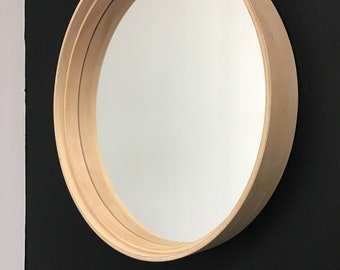 Op de afbeelding: Een ronde spiegel met een houten lijst met meerdere lagen hout die de spiegel omlijsten. De spiegel hangt aan een zwarte muur.