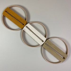May include: Three wooden hoops with string woven through them. The hoops are arranged in a row, with the top hoop having a mustard yellow string, the middle hoop having a white string, and the bottom hoop having a light brown string.