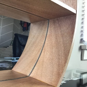 May include: A wooden shelf unit with a curved, rounded front edge. The shelf is made of light brown wood with a natural wood grain finish. The shelf has two shelves and is designed to be mounted on a wall.