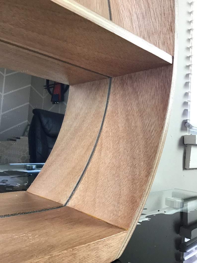 May include: A wooden shelf unit with a curved, rounded front edge. The shelf is made of light brown wood with a natural wood grain finish. The shelf has two shelves and is designed to be mounted on a wall.