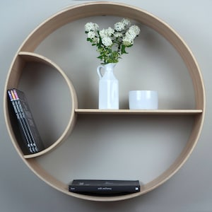 Puede incluir: Una estantería circular de madera con dos estantes. El estante superior tiene un jarrón blanco con flores blancas y un cuenco blanco. El estante inferior tiene un libro. La estantería está montada en una pared blanca.