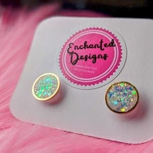 Puede incluir: Un par de pequeños pendientes redondos de purpurina iridiscente con engastes dorados. Los pendientes se muestran en una tarjeta blanca con el texto "Enchanted Designs Handcrafted Accessories".