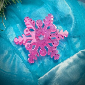 Puede incluir: Adorno de copo de nieve rosa lleno de purpurina. El copo de nieve está hecho de un material translúcido y tiene un diseño simétrico detallado. Descansa sobre un fondo de tela azul, con una ramita de vegetación en la esquina superior izquierda. Decoración festiva de invierno.