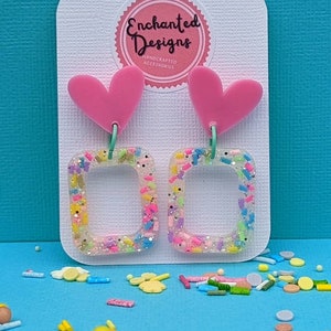 May include: A pair of earrings featuring pink heart-shaped tops and square frames filled with colourful sprinkles. The earrings are displayed on a white card with the text "Enchanted Designs". The background is a light blue surface.
