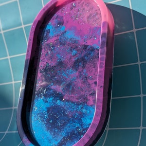 May include: A pink and blue resin tray with a galaxy-like design. The tray has a glossy finish and is decorated with glitter.