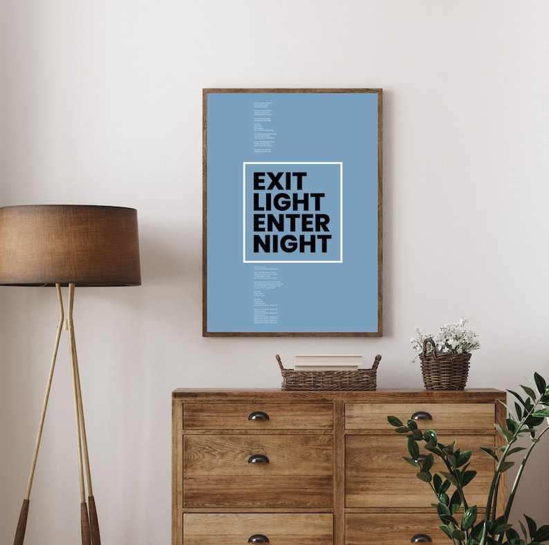 Op de afbeelding: Een lichtblauwe poster met de tekst "EXIT LIGHT ENTER NIGHT" in een witte doos op een houten dressoir met een lamp en planten.