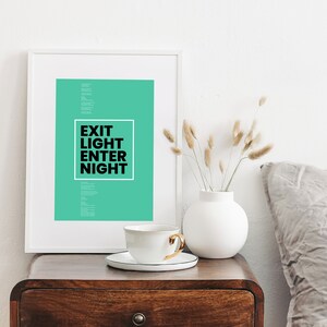 Op de afbeelding: Een witte ingelijste print met een teal achtergrond en zwarte tekst die "Exit Light Enter Night" luidt. De print staat op een houten tafel met een witte keramische vaas en een witte theekop.