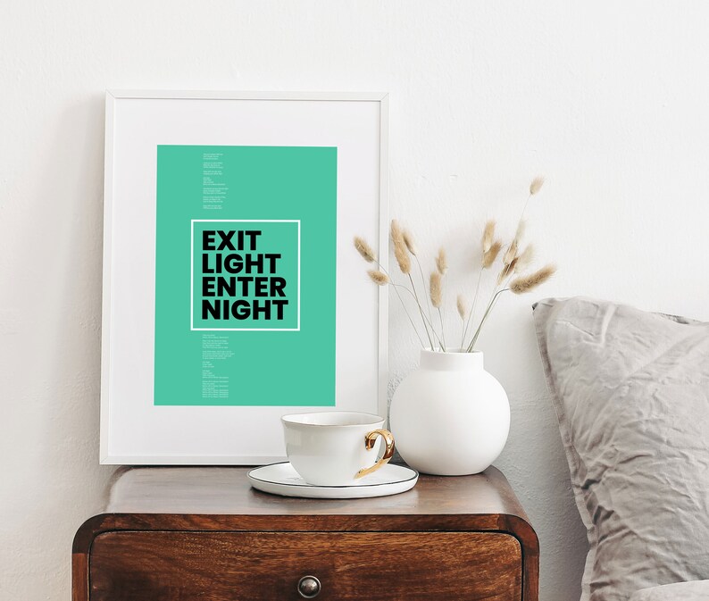 Op de afbeelding: Een witte ingelijste print met een teal achtergrond en zwarte tekst die "Exit Light Enter Night" luidt. De print staat op een houten tafel met een witte keramische vaas en een witte theekop.