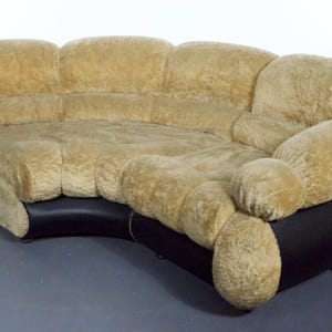 Może przedstawiać: Duża, zakrzywiona sofa narożna z miękkim, jasnobrązowym obiciem. Sofa ma unikalny, pikowany design i czarną skórzaną podstawę.