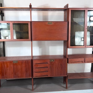 以下が含まれることがあります： ガラス扉と引き出しが付いた木製のミッドセンチュリーモダンなモジュール式壁ユニット。ユニットは暖色系のブラウン仕上げで、真鍮製の金具が特徴です。
