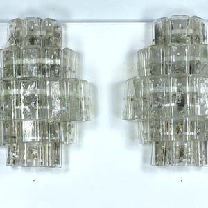 May include: A pair of clear glass wall sconces with a geometric design. Each sconce is made up of multiple layers of glass, creating a unique and modern look.
