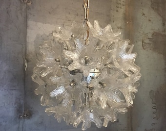 Puede incluir: Una lámpara de araña de cristal transparente con forma esférica y múltiples brazos con forma de flor. La lámpara de araña está suspendida de una cadena plateada.